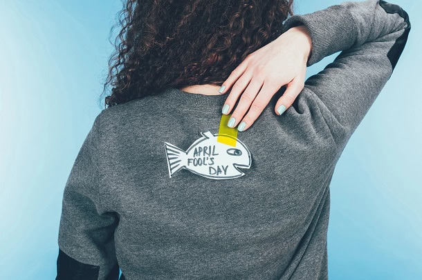 A woman trying to reach a paper sticker in a fish shape on the back on her sweater. The sticker says April Fools Day.