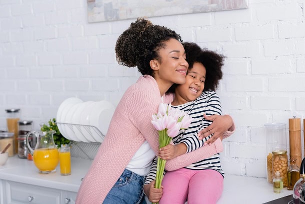 A mother hugging her daughter who is holding flowers.