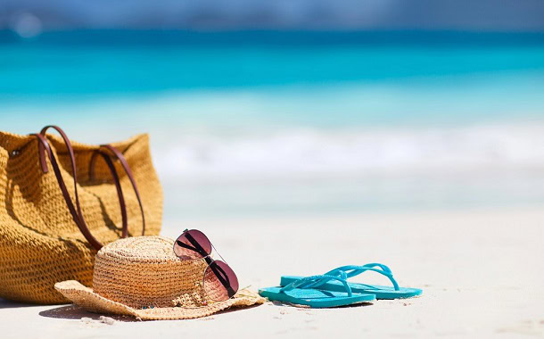 A beach with blue water and a bag, hat, sunglasses and a pair of flip flops.
