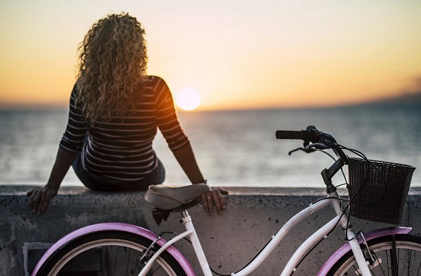 Woman looking at a sunset at the beach with her bike leaning at concrete in the foreground.