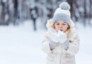 A small girl blowing at snow in her hands while standing in a forest in winter.