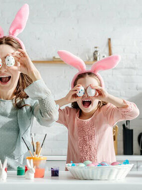 A happy mom and daughter in the kitchen holding up Easter eggs in front of their eyes.