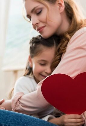 A daughter with a paper heart in her hand in the arms of her loving mother.