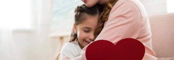 A daughter with a paper heart in her hand in the arms of her loving mother.