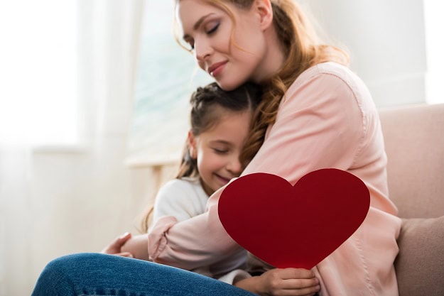 A daughter with a paper heart in her hand in the arms of her loving mother.