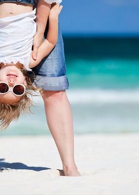 A small girl with sunglasses held upside down by an adult on the beach.