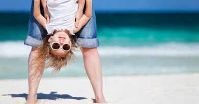 A small girl with sunglasses held upside down by an adult on the beach.