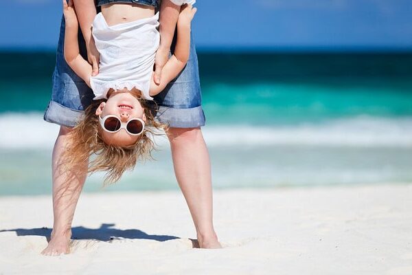 A small girl with sunglasses held upside down by an adult on the beach.