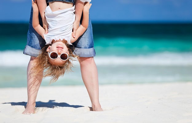 A small girl with sunglasses held upside down by an adult on the beach.
